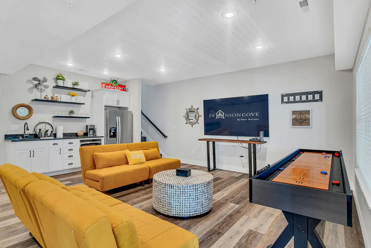 A cheerful, contemporary living area featuring a bold yellow sofa, a coffee table, a game table, and a large wall-mounted TV. The atmosphere is bright and energetic, perfect for socializing, playing games, or relaxing during the day. The yellow accents and modern decor add vibrancy to the space.