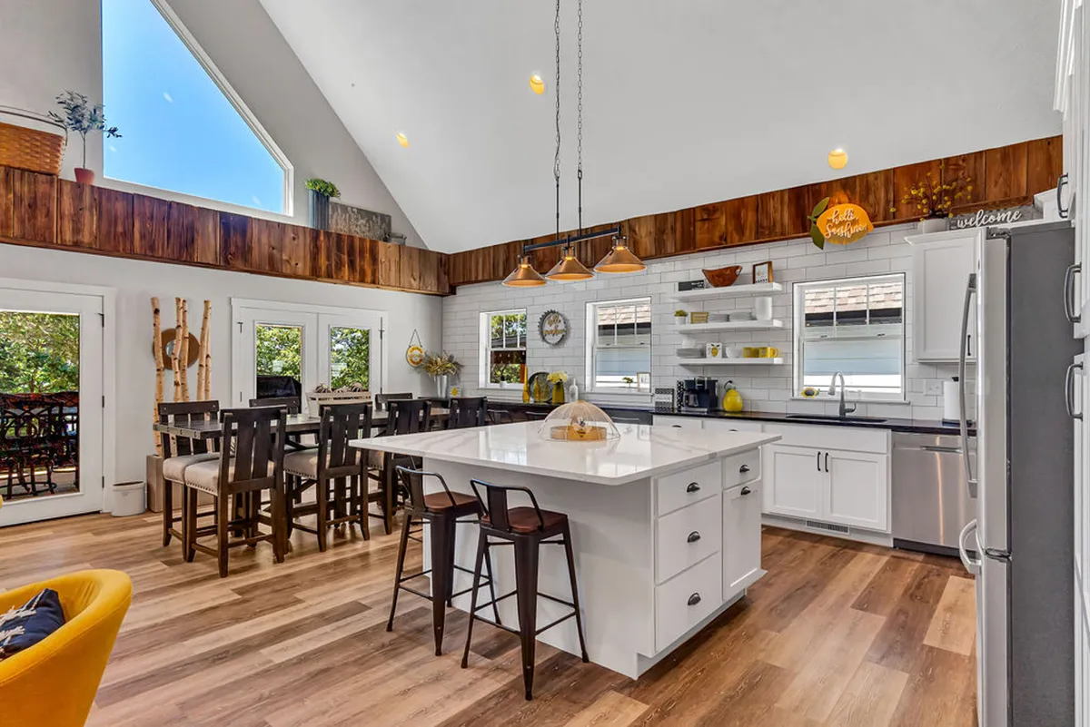 A spacious open-concept kitchen and dining area featuring a large marble kitchen island, exposed wooden beams, large windows bringing in natural light, and a generous dining table. Stainless steel appliances and stylish decor complement the gourmet setup, and the dining area seamlessly connects with the kitchen for group gatherings.