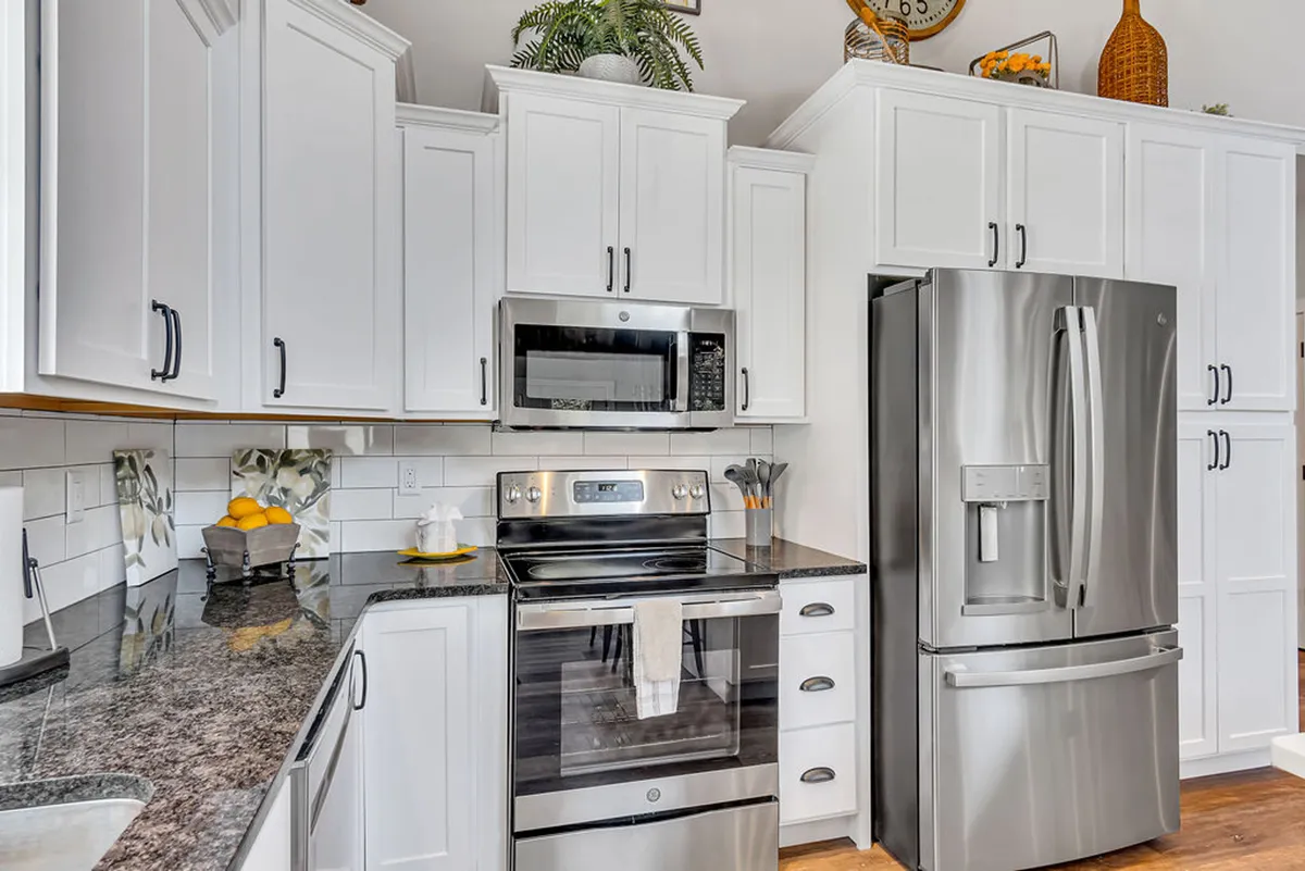A bright, airy kitchen with stainless steel appliances, a granite countertop, white cabinetry, and decorative touches like plants and fruit bowls. The layout encourages group cooking and socializing, while the finishes emphasize the gourmet, upgraded quality of the space.