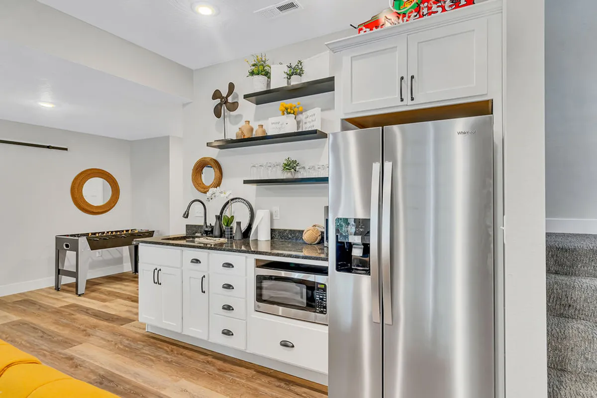 A multi-use entertainment area featuring a foosball table, round mirror, and a kitchen island with a stainless steel refrigerator. The space is open and bright, encouraging both active play and refreshments from the wet bar or fridge.