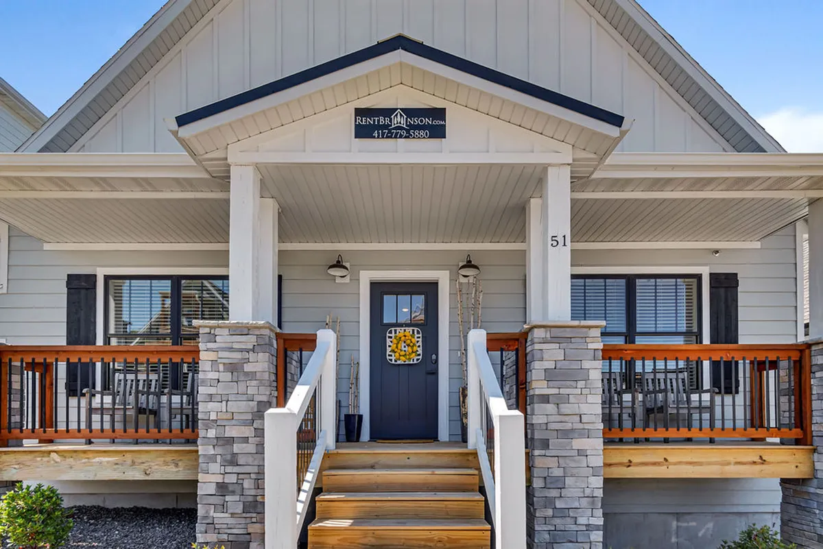 A welcoming front porch featuring stone pillars, wooden railings, and a decorative doormat, set against an inviting entryway. The porch is spacious, sheltered, and offers a sense of comfort and hospitality, ideal for relaxing with a pet.