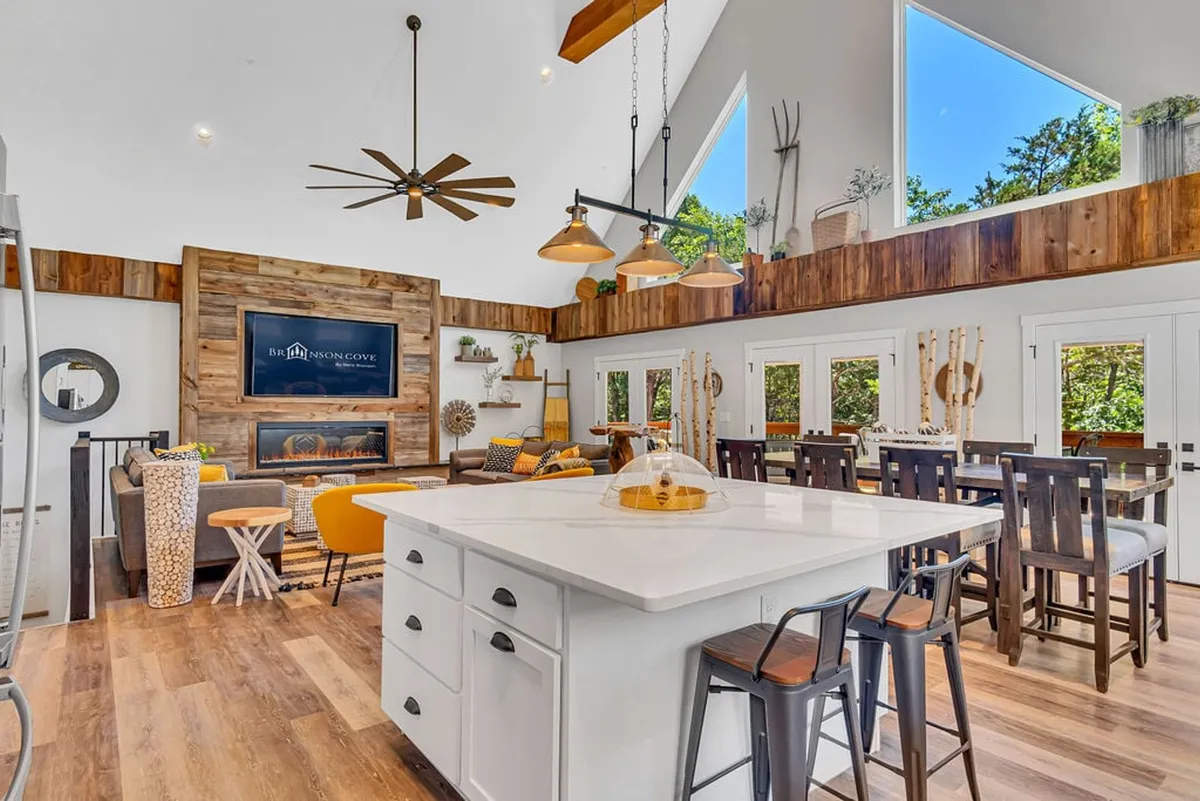 An expansive open-concept area blending the kitchen and living space, with a prominent fireplace, exposed wooden beams overhead, and large windows brightening the room. The seating and kitchen island area promote group gatherings and socializing, highlighting the room's versatility for both relaxation and entertaining.