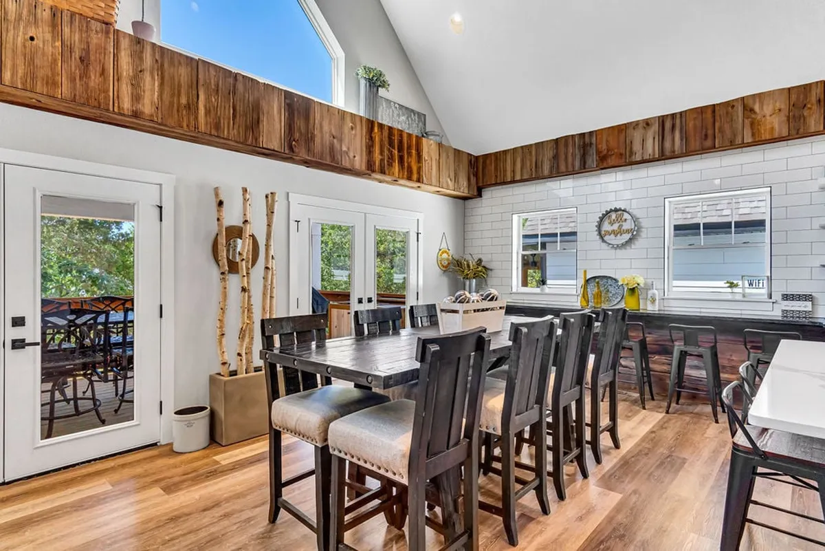 A warm, open-concept dining and kitchen area with a large dining table, wooden beams, abundant natural light from windows, and decorative touches. The setup invites large groups for meals, celebrations, and casual entertaining.